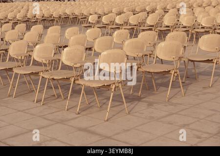 Vast rows of worn cream metal folding chairs arranged in a uniform grid on a stone plaza under warm golden hour light at an outdoor public event venue Stock Photo