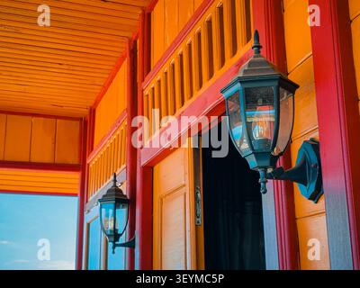 Bright  lantern hangs on wooden porch frame. Warm tones of wood contrast with deep red trim. Sunlit scene shows rustic charm and inviting atmosphere. Perfect for home decor, vacation branding Stock Photo