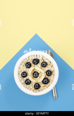 White bowl with overnight oats,bananas and blueberries on blue and yellow background Stock Photo
