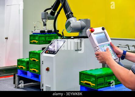 Industrial engineer operating automated robotic arm with teach pendant controller to lift green crates in modern factory warehouse Stock Photo