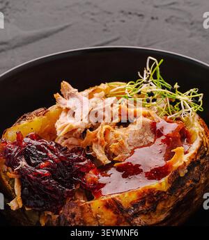 Delicious baked jacket potato loaded with tender pulled pork is served in black bowl. Nutritious comforting meal includes sweet caramelized onion chutney, savory barbecue sauce and fresh microgreens topping. Stock Photo