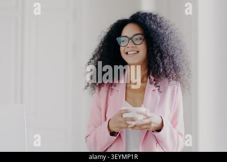 Photo of good looking African American prosperous busineswoman waits for partner in office, drinks coffee discusses future plans with colleague looks away with cheerful expression wears formal clothes Stock Photo