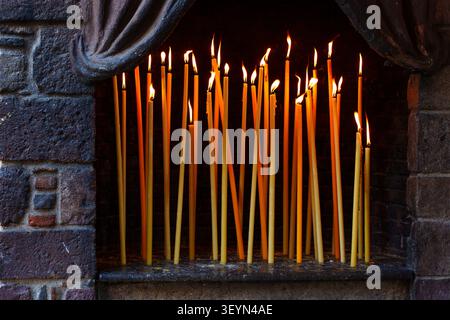 Glowing thin yellow candles burn brightly inside a rustic stone alcove, offering a spiritual and tranquil atmosphere at the Taxiarches Monastery in Le Stock Photo