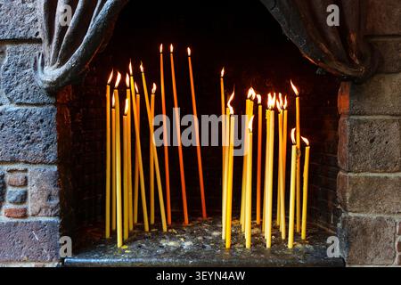 Slender yellow beeswax candles burn with bright flames inside a dark stone niche, creating a serene and spiritual atmosphere at the historic Taxiarche Stock Photo