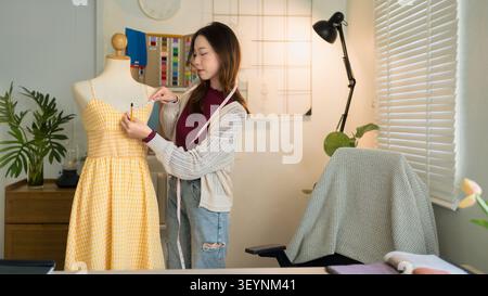 Young Asian woman fashion designer working on yellow gingham dress displayed on tailors mannequin in creative home Stock Photo