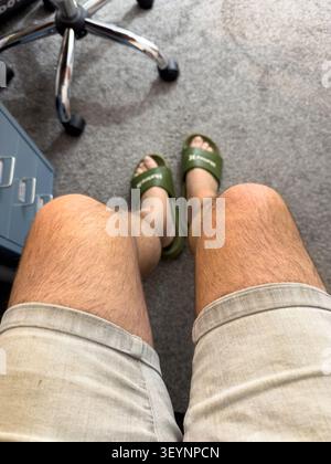 First-person perspective of a man wearing Hurley slides & denim shorts sitting indoors on a carpeted floor Stock Photo