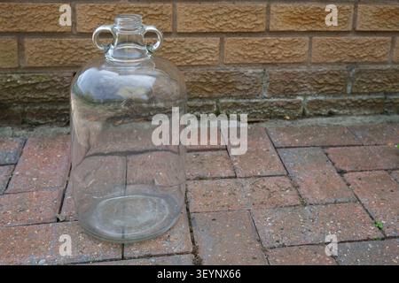 Single Standard Size One Gallon 4.5L Empty Clear Glass Demijohn On Red Brick Patio Stock Photo