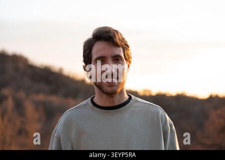 Young man smiling, looking at camera with happy expression. Golden hour light illuminating face, enjoying nature at sunset Stock Photo