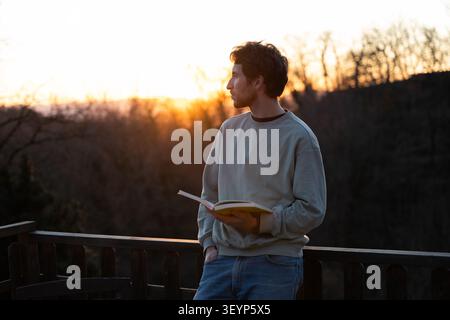 Young casual man standing on a balcony reading a book at sunset, enjoying a moment of contemplation, relaxation, and peace Stock Photo