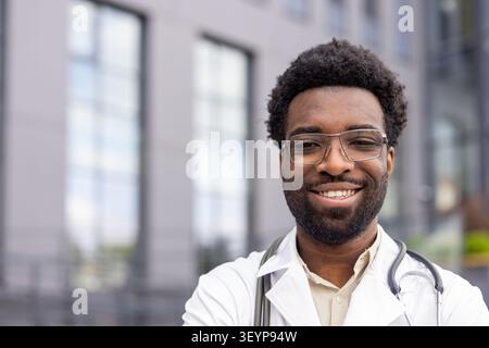 Male physician experiencing confidence and success in his career, standing outdoors against a modern building facade, symbolizing progress in urban healthcare and medical services Stock Photo