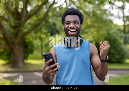 Cheerful young man wearing headphones and sportswear, clenching his fist in victory while looking at his mobile phone, celebrating achievement and fitness goals outdoors Stock Photo