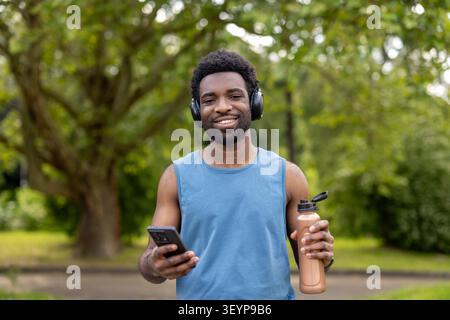 Young adult man listening to music with headphones, holding a smartphone and water bottle while smiling for the camera after a successful outdoor fitness workout in a green park Stock Photo