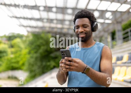 Young athletic person enjoying music through headphones and choosing songs on a mobile phone while standing casually outdoors at a sports arena Stock Photo