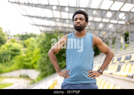 Focused man taking a moment of calm, catching his breath during a fitness workout, standing with hands on hips at an outdoor sports stadium Stock Photo