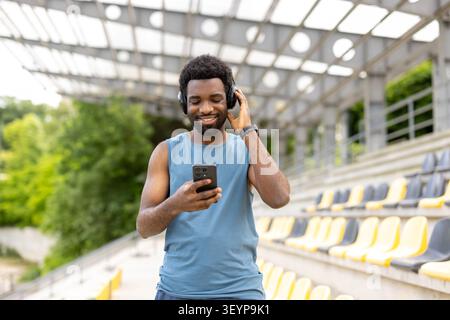 Young man wearing activewear and headphones, joyfully selecting music on a smartphone for an outdoor workout at a stadium, embracing technology for fitness motivation Stock Photo