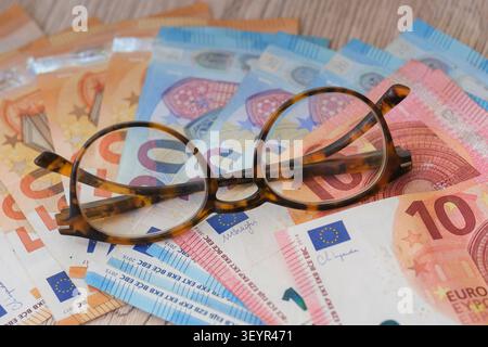 A pair of round reading glasses with a tortoise shell frame resting on a large fan of various Euro banknotes on a light wooden surface. Stock Photo