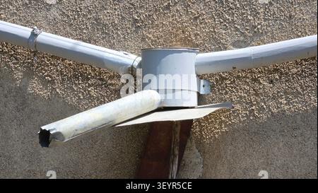 A round gray metal junction box links intersecting utility pipes with a specialized projecting spout on a rough stucco wall surface Stock Photo