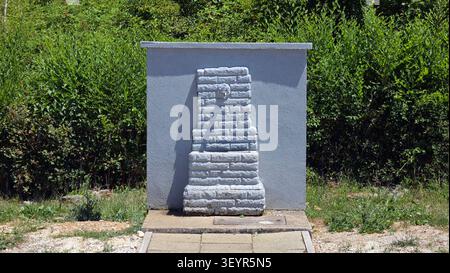 A light grey painted stone water fountain features a tiered brick texture pillar and a single metal spout centered against a flat concrete structural Stock Photo