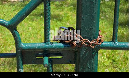 A close-up shot shows a weathered green metal gate wrapped in a brown rusty chain and locked tightly with a shiny brass padlock over a blurred green g Stock Photo