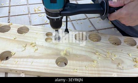 A close-up view of a person using a cordless drill to bore holes into a pine wood board. Stock Photo