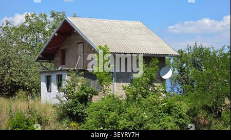 A modest multi-level countryside dwelling stands incomplete with exposed masonry walls and corrugated tiling under a clear blue sky Stock Photo