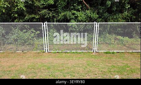 Three rectangular sections of a white painted steel lattice fence separate a dry grassy lawn from dense green bushes and forest foliage during the day Stock Photo