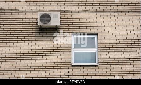 An HVAC compressor fan box hangs on a light tan masonry facade directly above a square double pane window with silver metallic framing under daylight Stock Photo