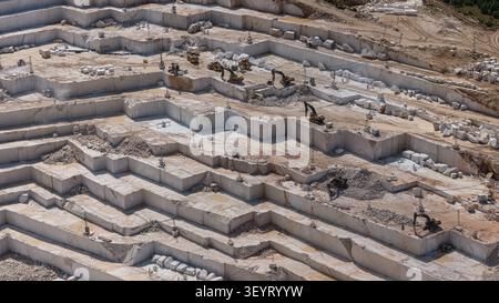 An aerial perspective shot of the massive terraced structure where marble blocks are cut in an open-pit mine, along with the heavy machinery. Stock Photo