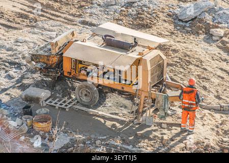 Worker in orange vest and red hard hat standing near mobile concrete pump, control panel open for maintenance, set against a typical construction envi Stock Photo