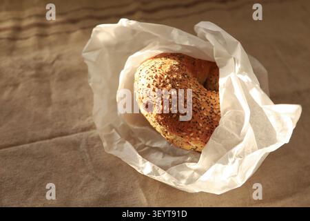 A golden brown bagel topped with sesame seeds sits inside a crumpled white paper bag on a textured beige linen fabric. Bright natural light creates so Stock Photo