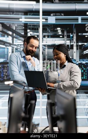 Cheerful team of engineers working with notebook computer in server environment, analyzing data. Stock Photo