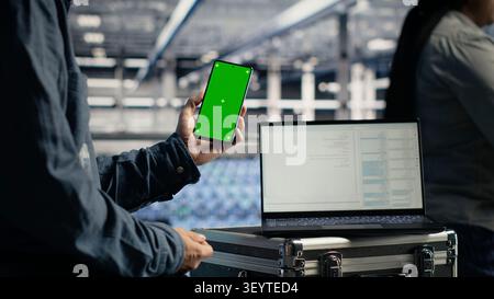 Software developer uses a green screen smartphone in a server room while waiting for code updates to finish. Stock Photo