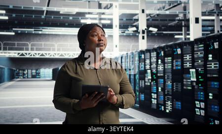 Admin walking in server facility, working with digital tablet to supervise software updates for system stability. Stock Photo