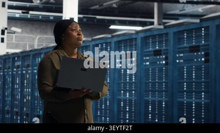 Cheerful systems engineer walking in server facility with notebook computer, checking hardware for system stability. Stock Photo