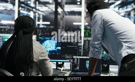 Server facility supervisor supervising staff member rolling out AI solutions optimizing equipment. Server environment supervisor supervising coder applying AI to minimize systems downtime. Stock Photo