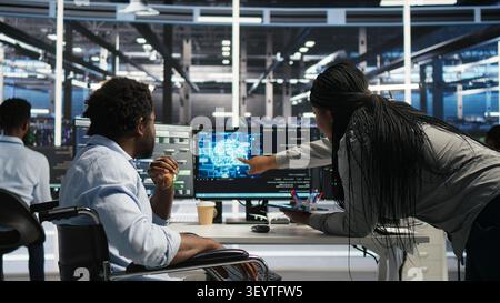Server room engineer in wheelchair and colleague oversee automated tasks working with node tree programming tool. Stock Photo