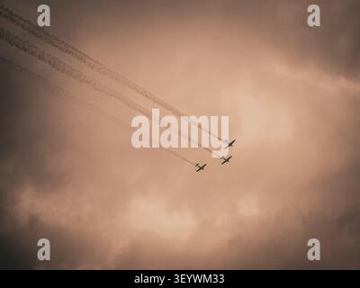 Three stunt airplanes flying in formation leaving long smoke trails across a dramatic sepia overcast storm sky during an airshow demonstration Stock Photo