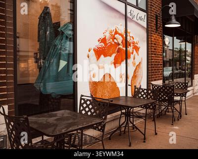 An empty outdoor cafe patio with black metal tables and chairs set against brick building facade featuring a large catering advertisement window sign Stock Photo