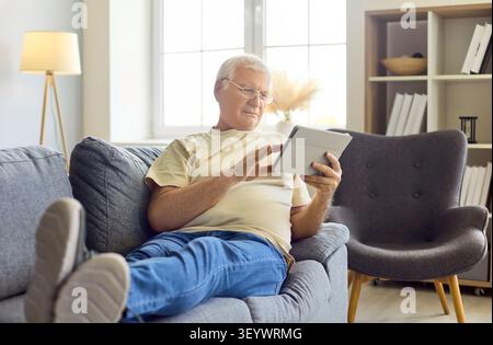 Senior man relaxing on sofa at home using tablet for leisure and browsing Stock Photo