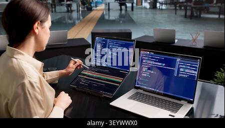 Young Female Software Developer Compares Code Changes On Dual Touchscreen Laptops. Stock Photo