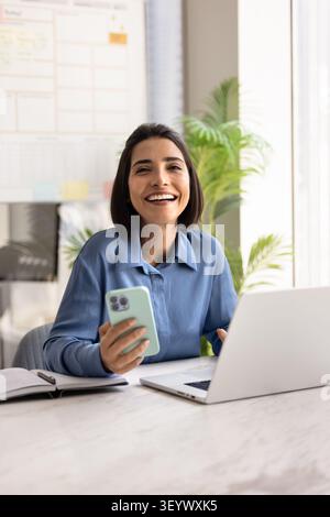 Smiling businesswoman using smartphone and laptop at workplace Stock Photo