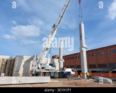 White mobile crane lifting a heavy precast concrete pillar while two construction workers guide it into place at a building site. Stock Photo