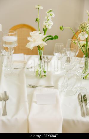 A wedding table setting with bud vase flowers, candles, and crystal glassware is arranged on white linen with rattan chairs Stock Photo