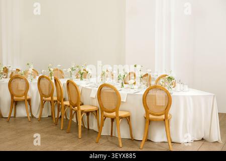 A long wedding reception table is set with white linens, rattan chairs, and delicate bud vase florals in a minimalist indoor venue Stock Photo