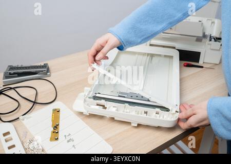 Hands carefully disassembling a flatbed scanner on a wooden desk, showing internal parts, cables and tools during repair and maintenance in a home off Stock Photo