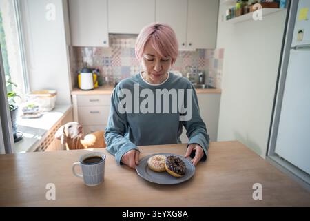 Mature woman sugar addict having lunch break with sweet chocolate doughnuts and cup of coffee. Stock Photo