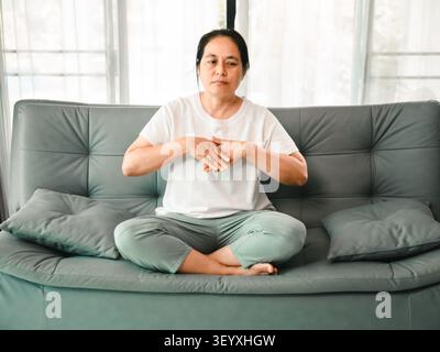 Middle aged Asian woman sitting cross legged on sofa and practicing breathing exercise at home, concept of wellness, meditation, stress relief, mindfu Stock Photo