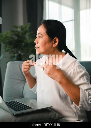 Middle aged Asian woman feeling hot and uncomfortable while working from home with laptop on sofa, adjusting white shirt collar in bright living room Stock Photo
