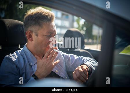 Stressed businessman smoking nervously while stuck in traffic, looking at his watch. Running late for work, urban commuting stress. Stock Photo