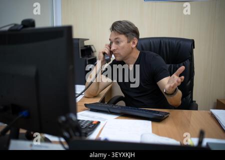 Focused man talking on landline phone while gesturing at his office desk. Business communication, negotiation, and active work. Stock Photo